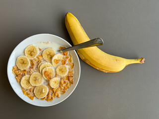 Bowl of cereals with sliced banana and a banana peel next to it on a grey table background viewed from above 