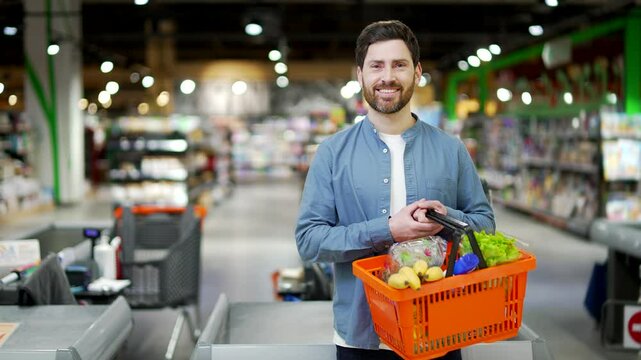 Portrait of a happy shopper in a grocery store or supermarket with a basket of groceries and purchases. Satisfied smiling customer handsome man looking at camera copy space