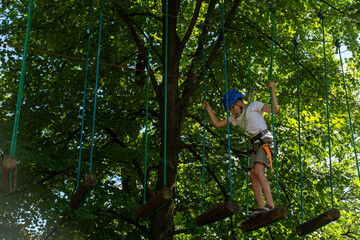 Daring teenager in a blue helmet navigates a challenging ropes course high above the ground in a dense forest. The concept of physical fitness and outdoor activities. Fear of heights. The psychology