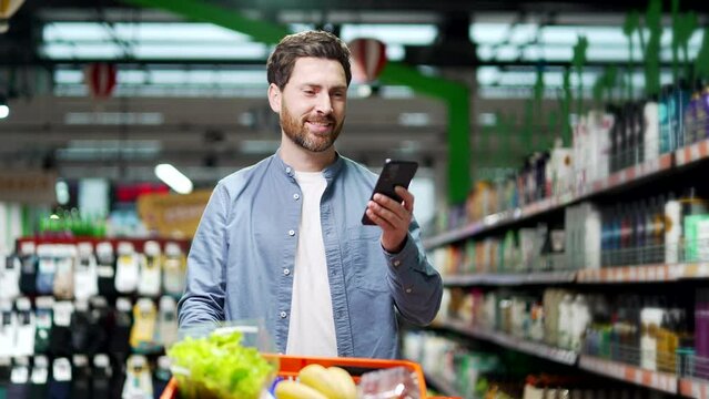 Handsome middle aged man buyer customer checking shopping list using smartphone, purchasing in grocery store in supermarket male shopper pick use mobile phone in hypermarket or food market deli