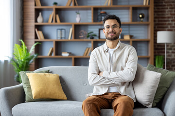 Obraz premium Smiling young Indian man sitting on sofa at home, arms crossed on chest and looking confidently at camera