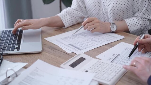 Close-up of a woman's hand checking financial paper beside a laptop on the wooden desk. Bookkeeping, finance audit and taxes. Business concept