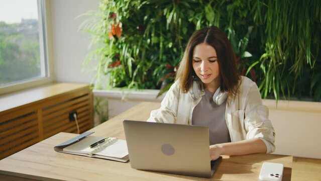 In the well lit office, a woman creates a productive workspace with greenery and modern technology at her wooden desk. The calm and efficient environment boosts her focus and concentration