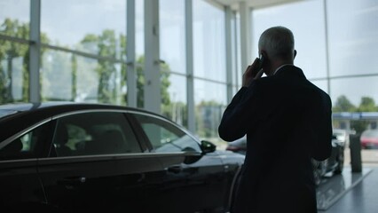 Back view of busy man in business suit standing in shadow office. Businessman remotely talking on smartphone in large showroom. In middle of auto shop located luxury cars. Dealership concept. - Powered by Adobe