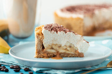 Slice of banoffe pie filled with banana and dulce de leche and sprinkled with grated chocolate. Banana cake on white plate with glass of coffee on turquoise summer background