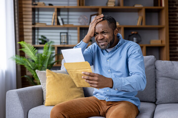 A middle-aged man seated on a living room couch reading a letter with a distressed and worried...