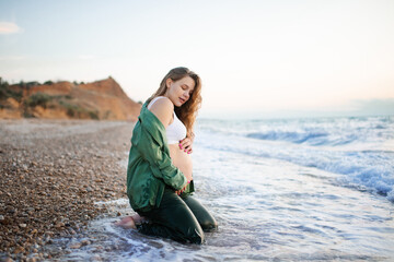 Pregnant woman 25 - 29 year old wearing silky clothes holding naked tummy over sea shore outdoor. Maternity lifestyle. Motherhood.