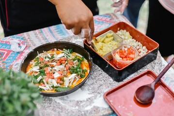 A person is putting toppings on a pizza in a kitchen. The kitchen countertop has a checkered tablecloth. There are pieces of pineapple, diced tomatoes, and chopped onion on the dishes near the pizza.