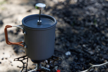 A French press coffee maker sits atop a small camping stove, nestled amongst cooled embers, ready to brew the perfect morning cup.