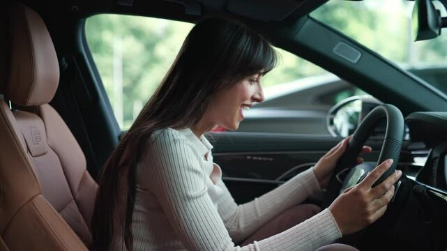 Side view of happy Caucasian woman enjoying test driving new automobile. Beautiful client happily observing equipment in car salon. Touching steering wheel with hands. Selecting best auto.