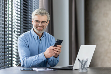 Confident mature businessman sitting at his desk in a modern office, holding a smartphone, and smiling at the camera. He is working on his laptop and looks happy and approachable