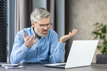 Senior man in a blue shirt expressing frustration while working on a laptop at home. The man...