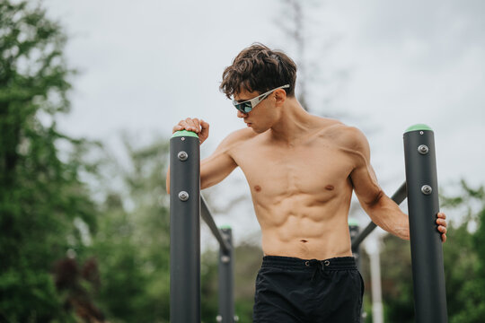Athletic young man showing strength and fitness while working out on parallel bars outside in a green park.