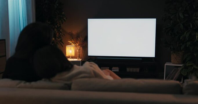 Happy young girls women night evening. Two friends sitting in the living room on sofa gouch relaxing watching tv movie films tv with white screen mockup at cozy home.