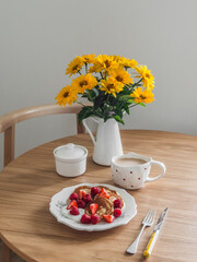 An aesthetic morning - pancakes with fresh berries, coffee, a bouquet of flowers on a round wooden table
