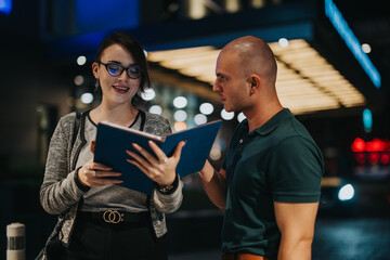 Two business professionals are discussing work at night in an urban environment, holding a blue folder.