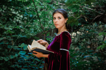 A young woman with brown hair in a medieval dress with a book in her hands stands in the forest