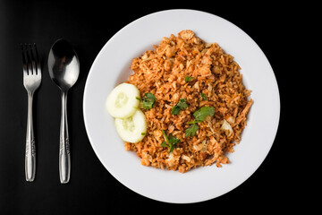 Close up flat lay photo of Homemade fried rice with vegetables, chicken and fried eggs served on a white plate with spoon and fork. Isolated on black background.