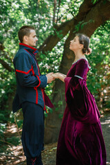 A man and a woman in 19th century costumes in a forest. The couple look at each other and hold hands