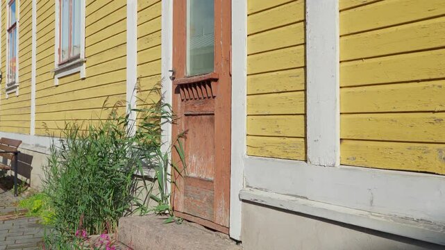 Yellow Wooden House Exterior in Pori, Finland	A yellow wooden house with white trim stands in Pori, Finland. The house has a single wooden door with a glass window, surrounded by green foliage. A ston