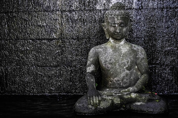 Obraz premium Stone statue of a Buddha figure meditating facing the front with temple stone wall background. Bodhisattva Face. Concept for Vesak Day and Enlightenment Day. Empty blank copy text space