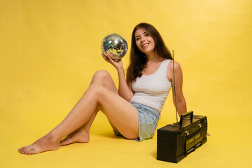 A young woman in denim shorts and a T-shirt sits on a yellow background and holds a disco ball in her hand. There is a retro tape recorder nearby