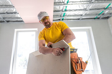 The worker make a plasterboard ceiling. He doing adjusts a size of the board using knife cutter. A...