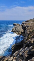 Dramatic coastal landscape. Rugged rocky cliffs meet deep blue ocean. Crashing waves, white foam. Clean sky. Natural scenery. Seascape. Shoreline. Rocky shore. Outdoor nature photo.