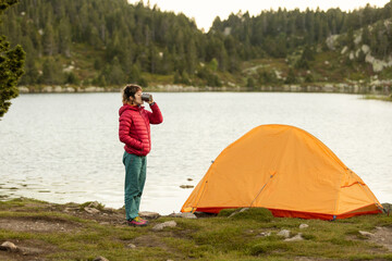 A woman is drinking coffee by a lake in front of a tent. The scene is peaceful and serene, with the woman enjoying the natural surroundings