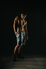 Athletic young man posing shirtless in dark studio, highlighting his muscular body with dramatic lighting for fitness and strength concept.