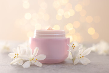 Cream in jar and jasmine flowers on grey table against beige background with blurred lights, closeup