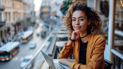 Fototapeta premium A lady with curly hair works on her laptop at an outdoor cafe with a scenic view of the city, embodying the modern balanced lifestyle of work and relaxation.