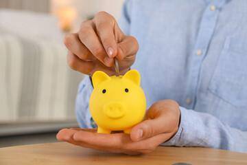 Man putting coin into yellow piggy bank on wooden table, closeup