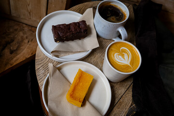 Coffee break. Two mugs of American and latte art on a wooden background. Chocolate brownie and yuzu cake slices. Delicious sweet food and coffees. Flat white, latte. Food and drink.