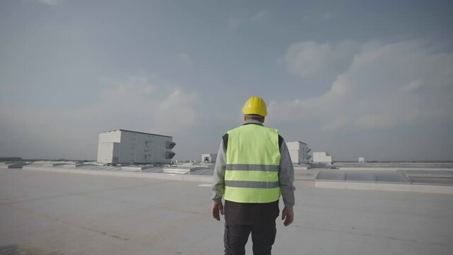 Back view of man wearing safety equipment walking on flat roof of modern commercial or industrial building. Forwards tracking on engineer.