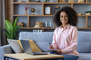 Smiling woman in a pink shirt taking notes while working on a laptop from home. Cozy home office setup with bookshelves in the background. Perfect image for remote work, studying, or home