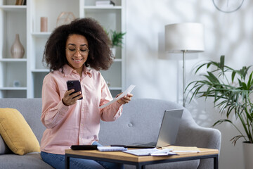 Happy woman managing her finances at home, using a smartphone to check her expenses while holding a...