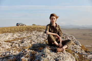 Adventurous woman enjoying serene view of mountains while resting on rock near car in nature landscape