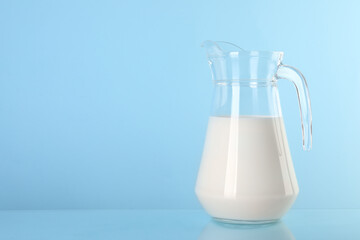 Glass jug of fresh milk on light blue background, space for text