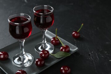 Delicious cherry liqueur in glasses and berries on black table, closeup. Space for text