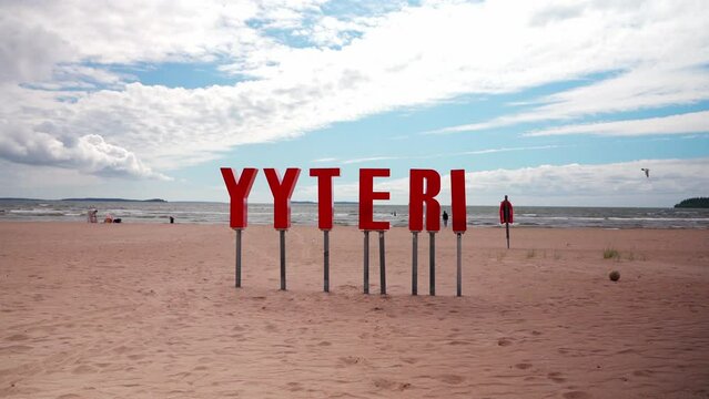 A red sign spelling Yyteri stands on a sandy beach, with the Gulf of Bothnia in the background. A few people walk along the beach in the distance, while the sky is a bright blue with fluffy white clou