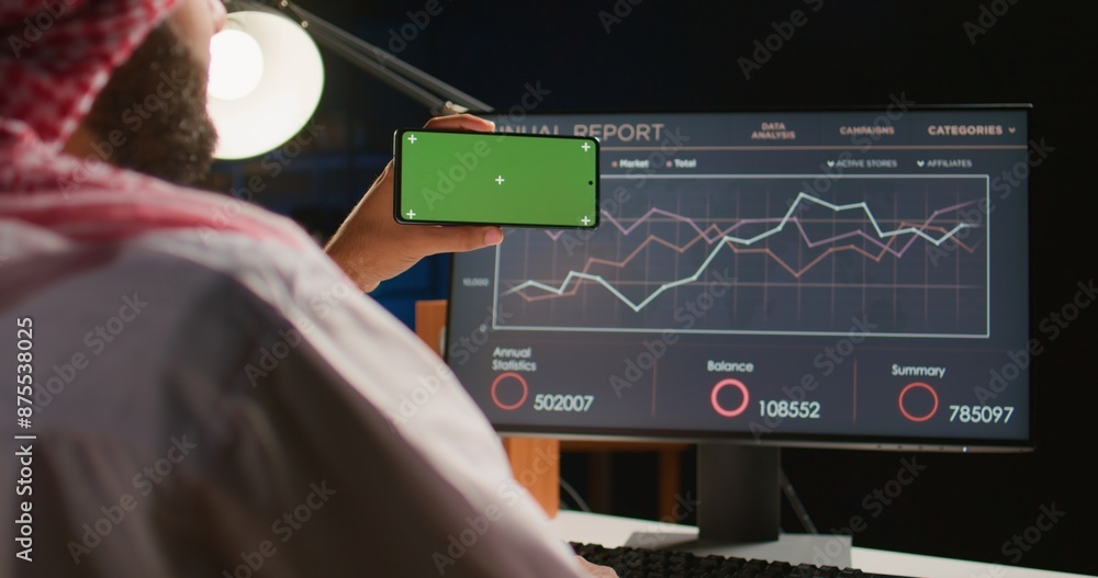 Poster man holding green screen cellphone while looking at business annual report charts on computer. self 