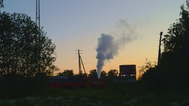 Smoke coming from diesel locomotive in golden twilight. Dark silhouettes of trees and electric poles on railway station. Transportation of heavy shipment by coal loco train using railroad. Transport