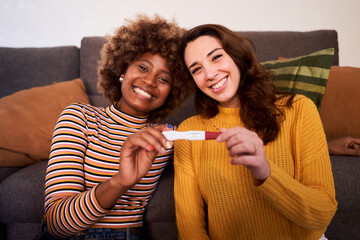 Cheerful portrait of expecting lesbian multicultural couple at home showing the positive pregnancy test and looking at camera happily. © Gigi Delgado