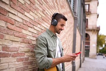 Attractive young Caucasian man with wireless headphones using cell phone leaning against brick...
