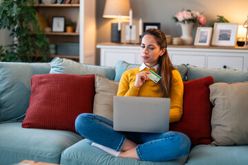 Young woman looking suspicious at laptop screen while holding credit card and sitting on a couch at...