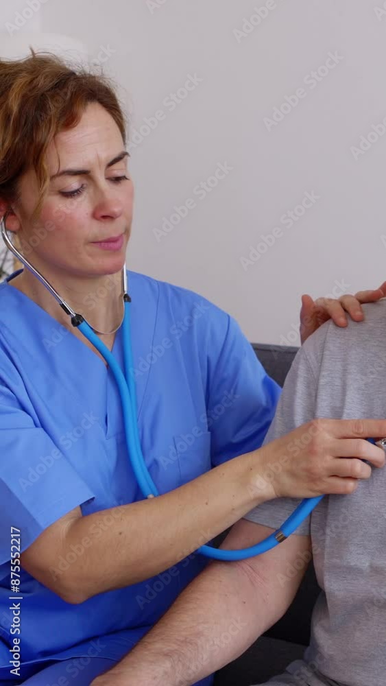 Wall mural Vertical HD video of young female doctor in blue uniform listening to disabled older patient heart sound with stethoscope during visit at home. Medical checkup and healthcare concept