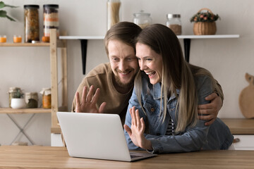 Obraz premium Joyful young 35s spouses spend time in the kitchen, using videoconference application, make videocall chat with friends, greet family, smile, enjoy virtual meeting at home. Teleconference, remote talk