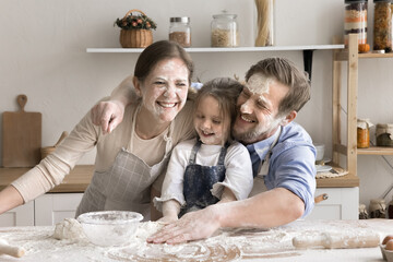 Playful parents and little daughter having fun while preparing dough for homemade pie or cake, they faces smeared with white flour, family laughing, fooling feel happy spend time together in kitchen