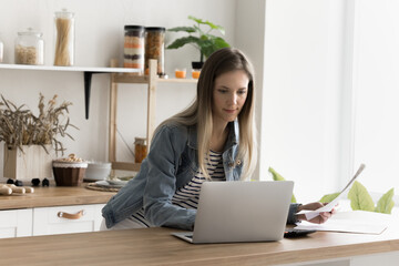 Young focused woman stands in home kitchen, do accounting work, hold paper document, check household bills, pay utility use modern bank application on laptop, busy in paperwork, comparing information